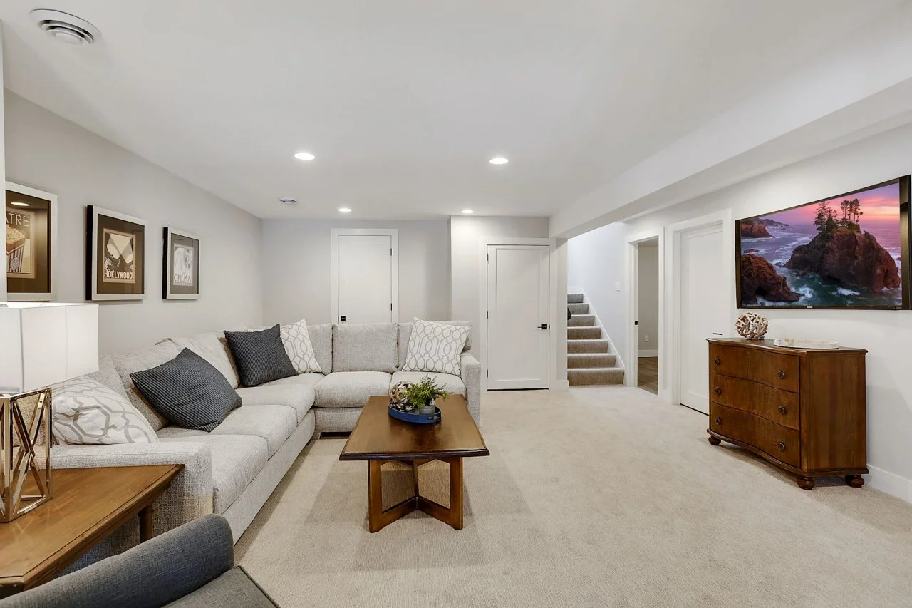 A beautifully finished basement with a modern, cozy design featuring a large L-shaped light gray sectional sofa, a wooden coffee table, and a TV mounted on the wall. The space has recessed lighting, carpeted flooring, and neutral-colored walls. A wooden dresser sits against one wall, and framed artwork decorates another. A staircase leads to the upper floor. The image represents a well-renovated basement, illustrating potential basement finishing costs for homeowners considering similar upgrades.